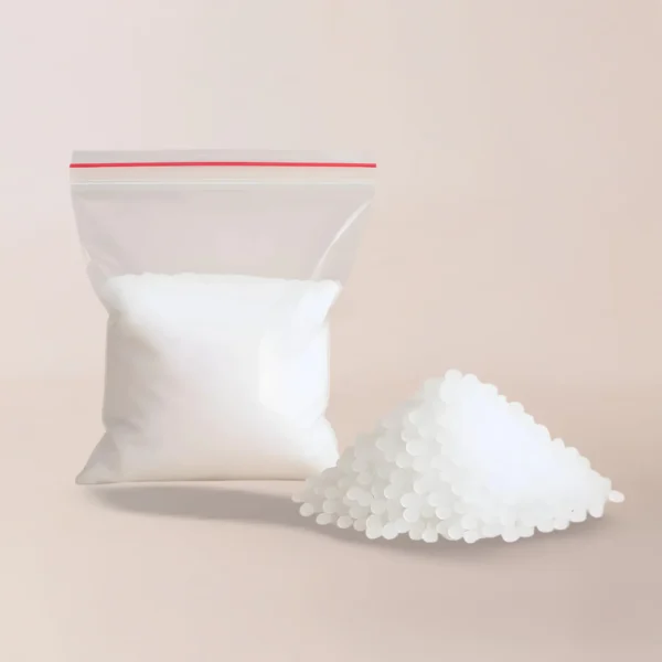 Una bolsa de plástico transparente etiquetada como Aditivo Vybar se encuentra en posición vertical junto a una pila de bolitas blancas sueltas sobre una superficie de color beige claro, ampliamente utilizada como ingrediente para velas por fabricantes de velas y entusiastas de la fabricación de velas.