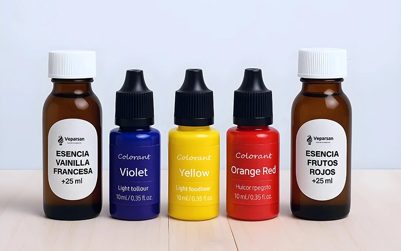 Cinco frascos de colorante alimentario y esencia, perfectos como insumos para velas, están dispuestos en fila; las etiquetas indican vainilla, violeta, amarillo, rojo anaranjado y fruto rojo, cada uno con volúmenes específicos.